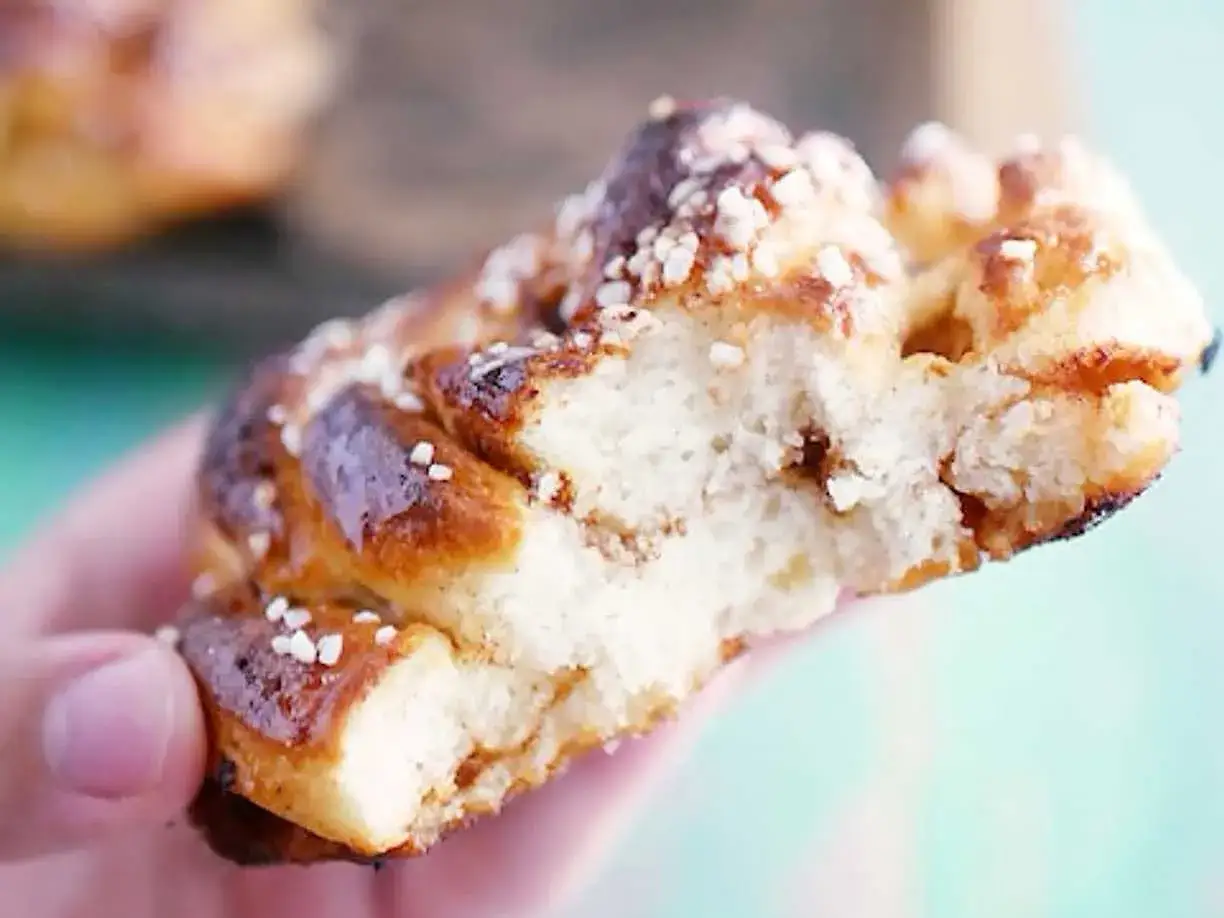 En hand håller en bit av en gyllene, nybakad glutenfri kanelbulle, strödd med pärlsocker.