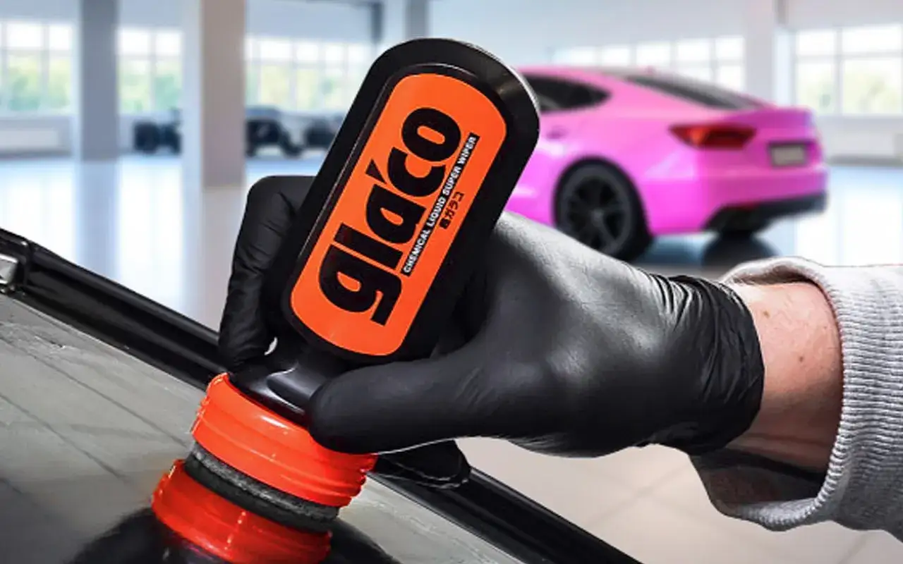 Hand med handske applicerar glaco glasförsegling på en bilruta. En rosa bil syns i bakgrunden.