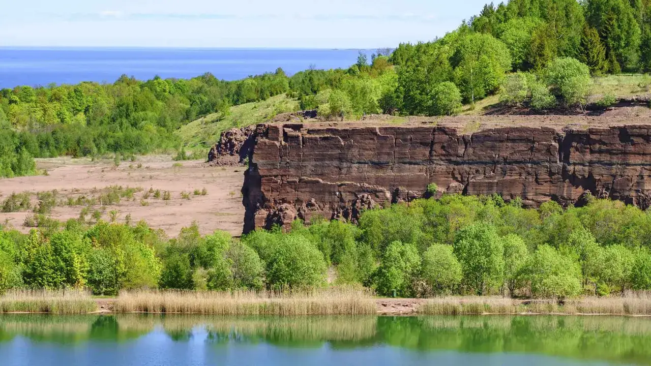 Vacker utsikt &ouml;ver en sj&ouml; med vass, en brant klippa och gr&ouml;nskande tr&auml;d. Perfekt f&ouml;r en kinnekulle vandring.