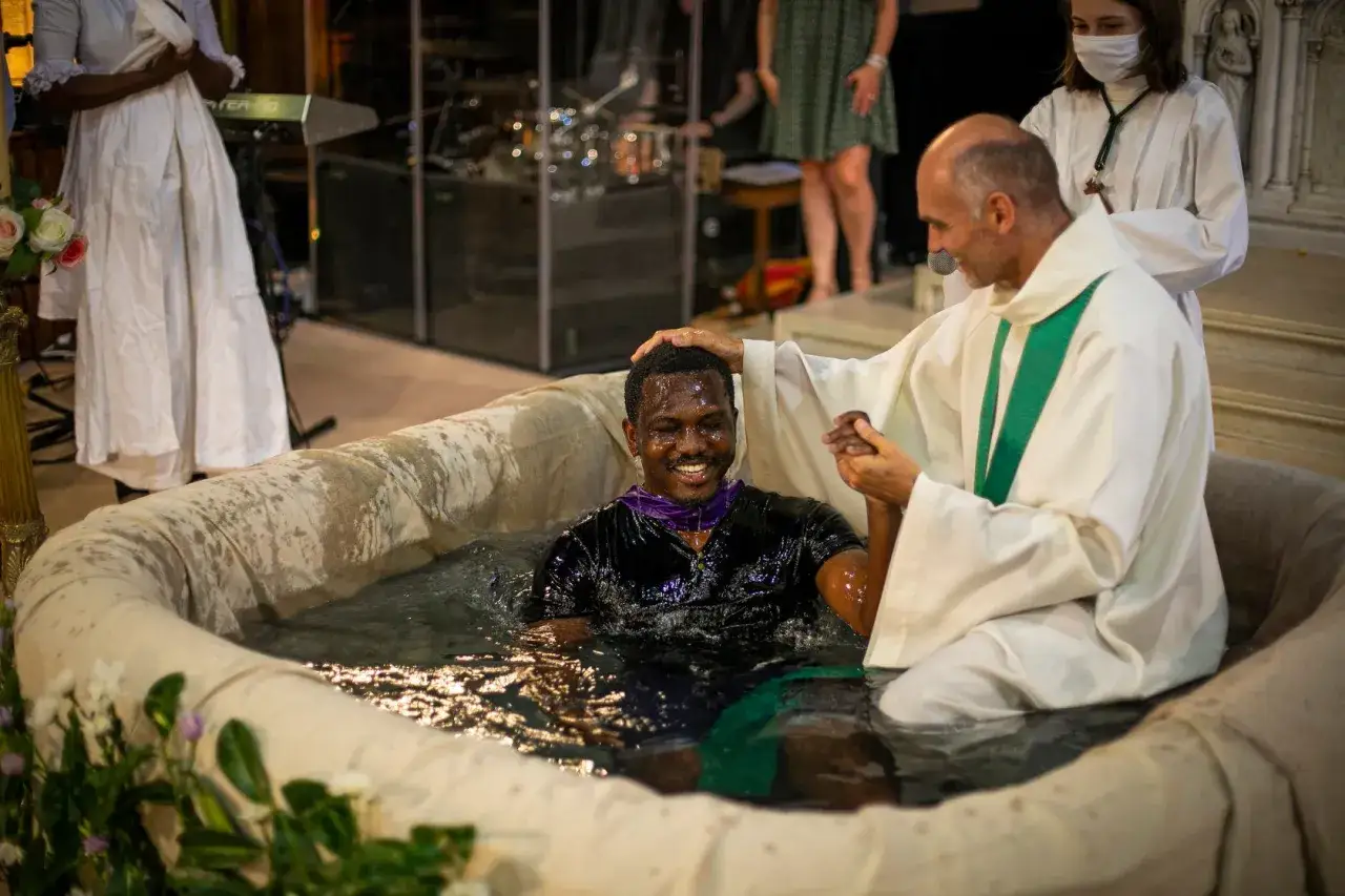 Un homme souriant est immergé dans l'eau d'une cuve baptismale. Un prêtre, en robe blanche, lui pose la main sur la tête. C'est qu'est ce que le baptême catholique.
