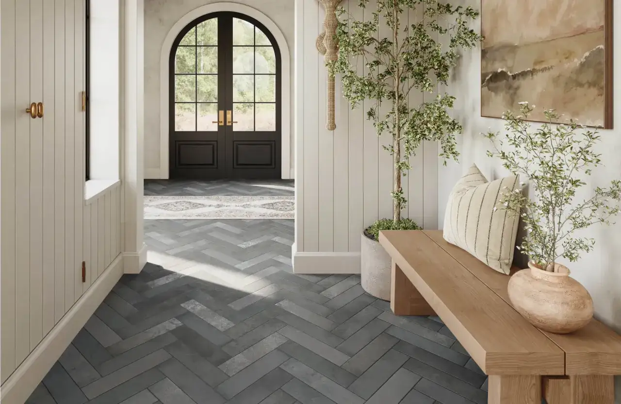 Un vestibule lumineux avec un sol en **carrelage en quinconce** gris, une porte cintr&eacute;e noire, un banc en bois et une plante verte.
