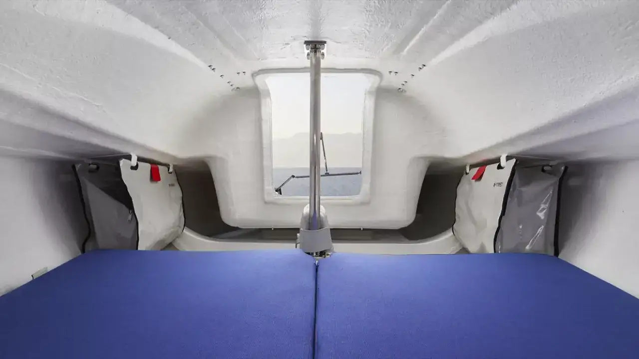 Intérieur d'un voilier avec matelas bleu, vue sur la mer par une fenêtre. Sacs de rangement suspendus. first 18 avis.