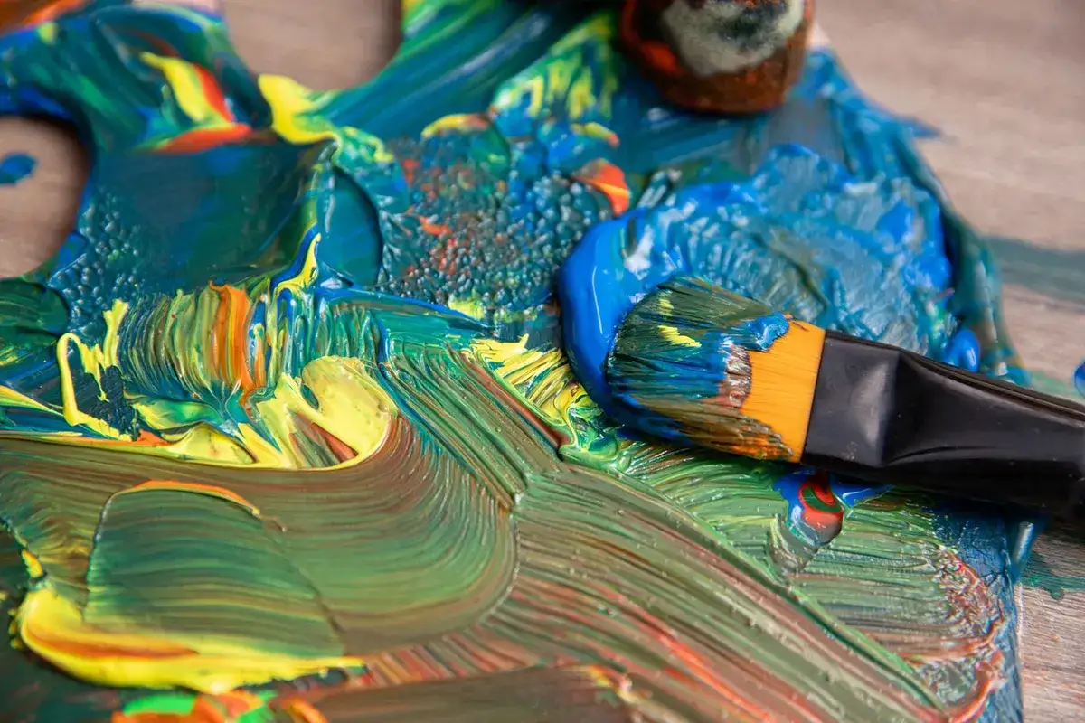 Un pinceau chargé de peinture bleue s'apprête à rejoindre une palette vibrante de verts, jaunes et oranges. L'œuvre d'un artiste peintre amateur prend forme.