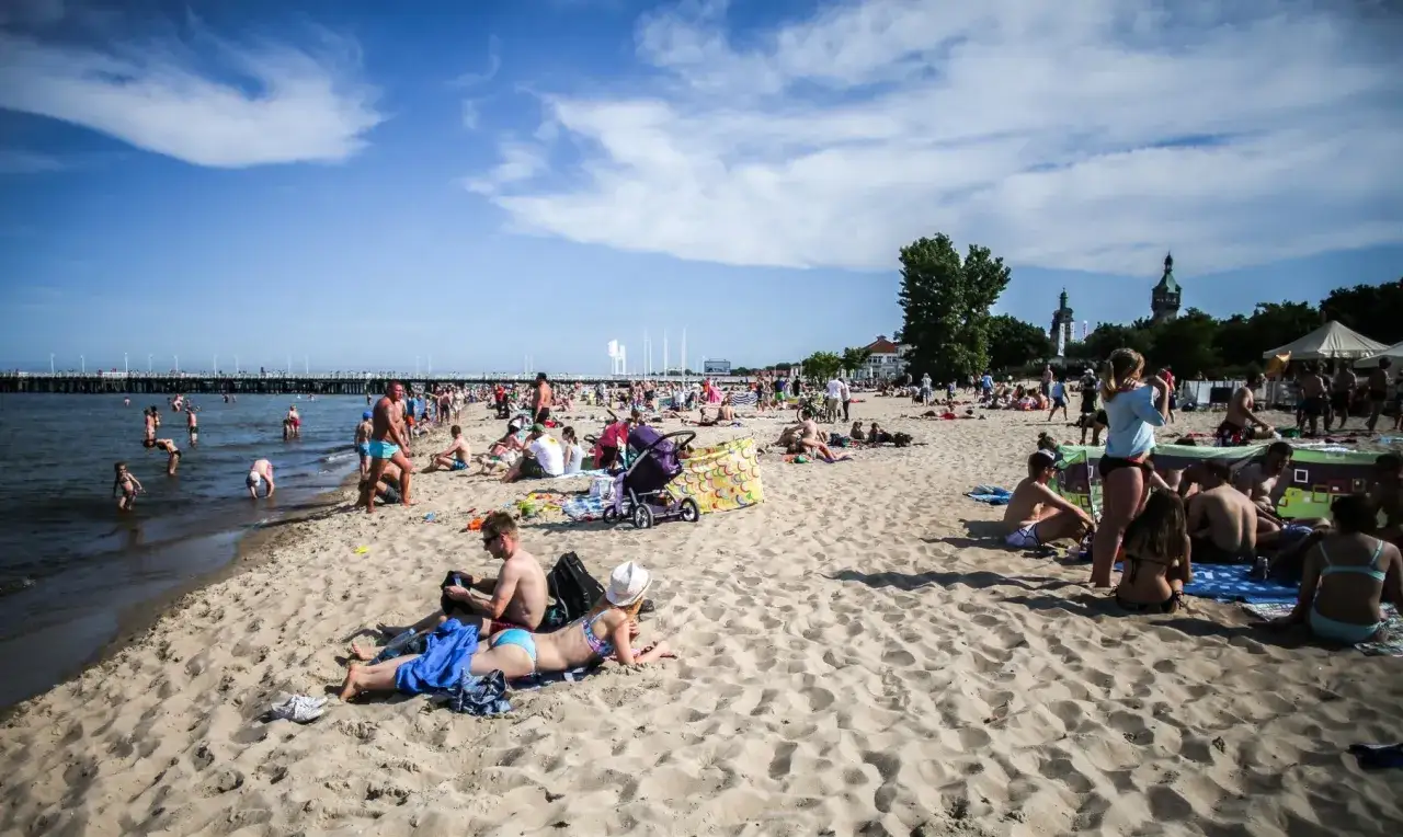 Tłum ludzi na plaży w Gąskach, cieszący się słońcem i morzem. Wiele rodzin z dziećmi, wózek dziecięcy i kolorowe parawany świadczą o tym, że to miejsce oferuje świetne gąski atrakcje dla dzieci.