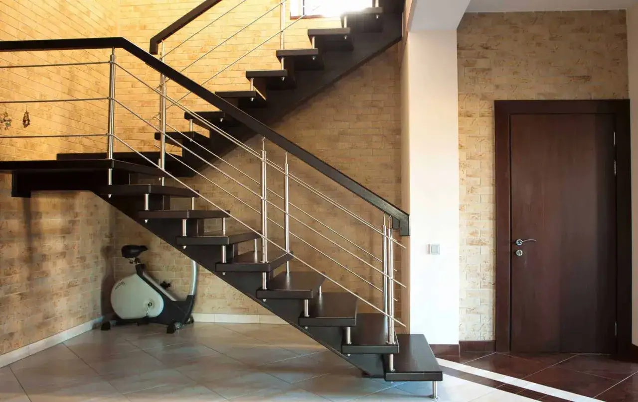 Nowoczesne schody w salonie z ciemnego drewna i metalową balustradą. Pod schodami stoi rowerek stacjonarny.