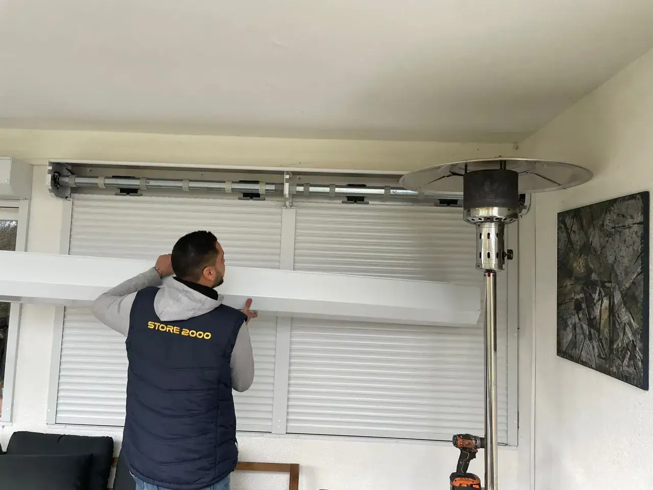 Un technicien installe un syst&egrave;me pour motoriser des volets roulants. Un chauffage d'ext&eacute;rieur est visible &agrave; droite.