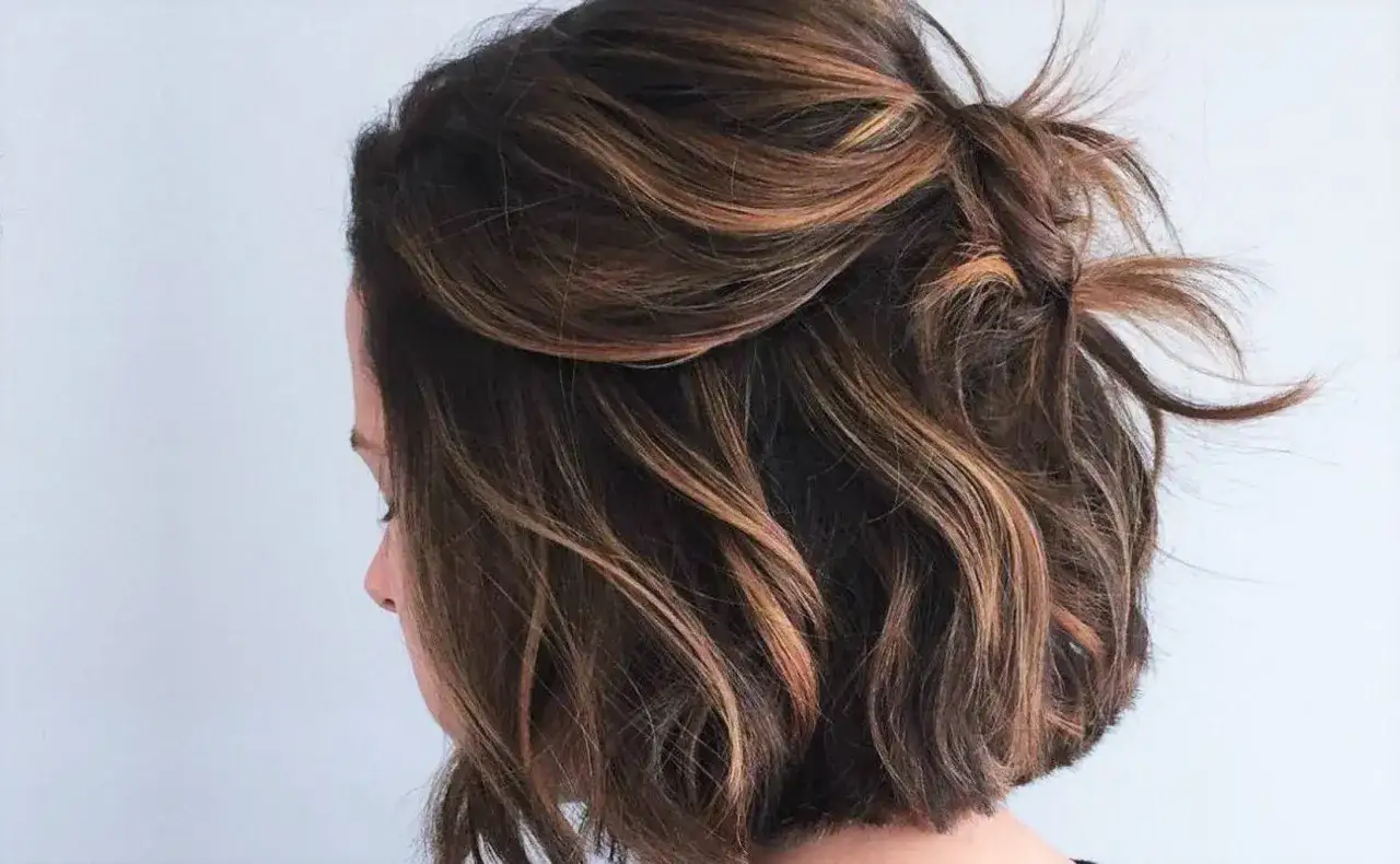 Chevelure brune avec des mèches subtiles sur des cheveux très courts, coiffés en un chignon flou et texturé.