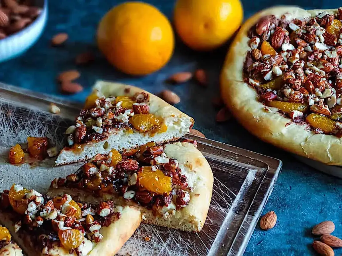 Süße Nutella Pizza mit Mandeln und Orangenstücken, auf einem Holzbrett serviert.