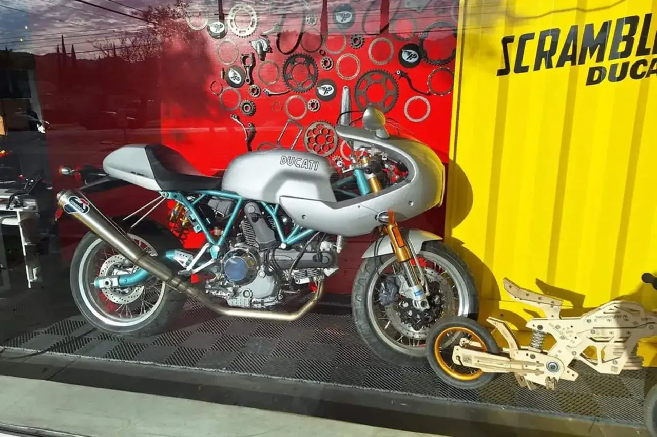 Ducati plateada estilo café racer, con detalles en azul y un escape Termignoni. A su lado, un modelo de madera de moto, quizás un homenaje a Paul Smart.