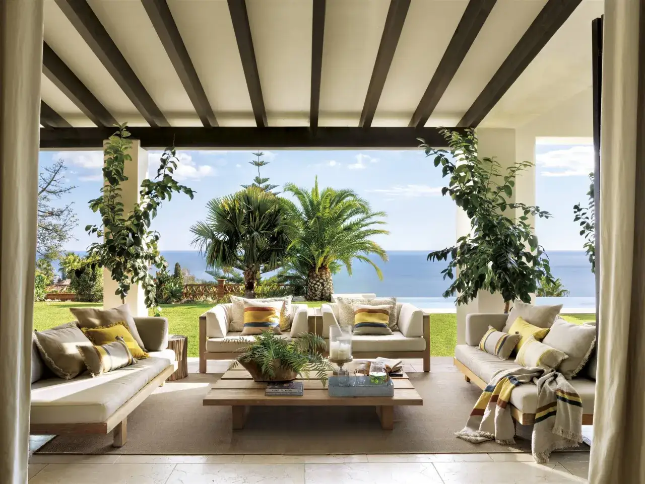 C&oacute;modos porches de entrada principal con sof&aacute;s de madera, cojines y mantas, con vistas al mar y palmeras.