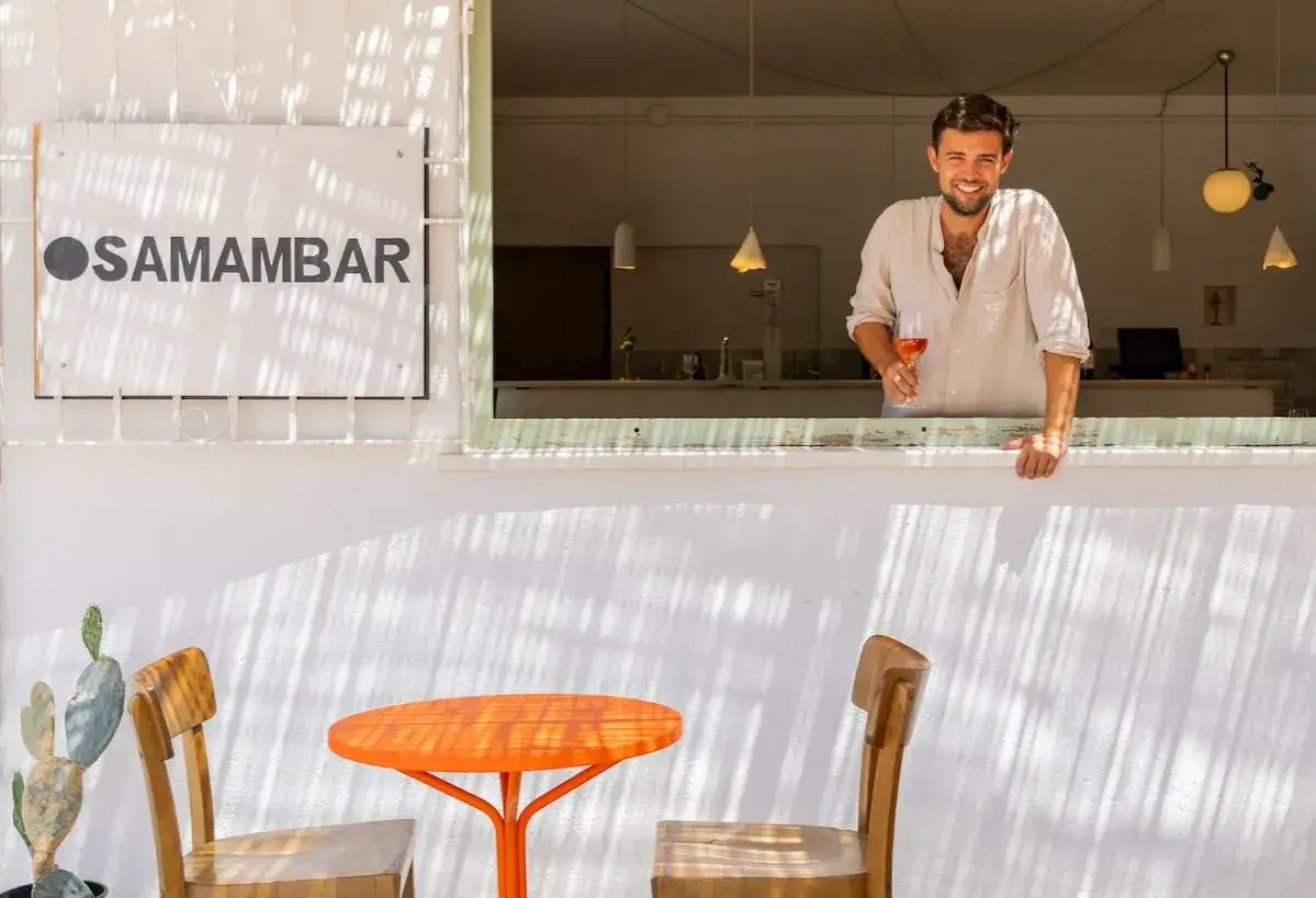 Hombre sonriente con copa de vino rosado en Samambar, ideal para comer en Rodalquilar. Mesa naranja y sillas de madera invitan a disfrutar.