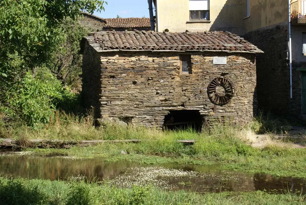 Antiguo molino de agua de piedra, con su rueda oxidada y el río tranquilo reflejando el verde.