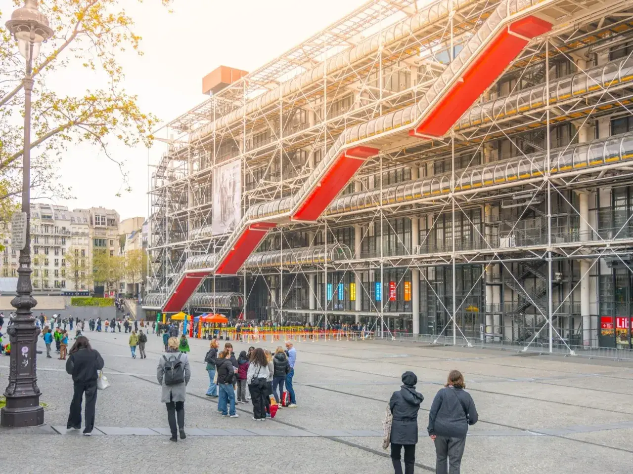 Widok na Centre Pompidou z czerwonymi schodami ruchomymi i ludźmi spacerującymi po placu.