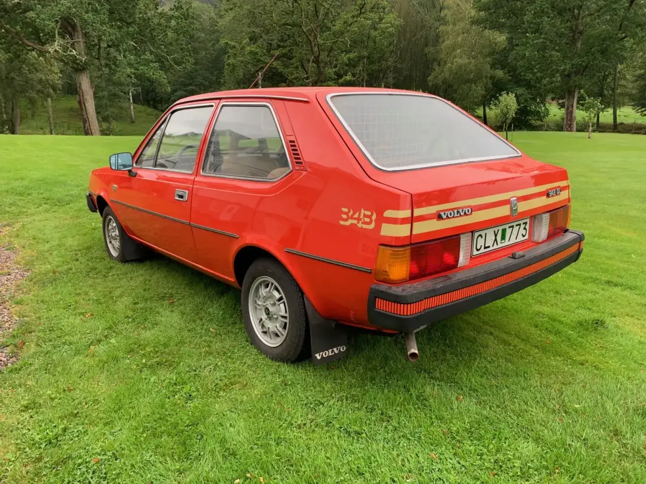En röd Volvo 343 står på en grön gräsmatta med skog i bakgrunden. Bilen har en guldfärgad rand längs sidan och siffrorna