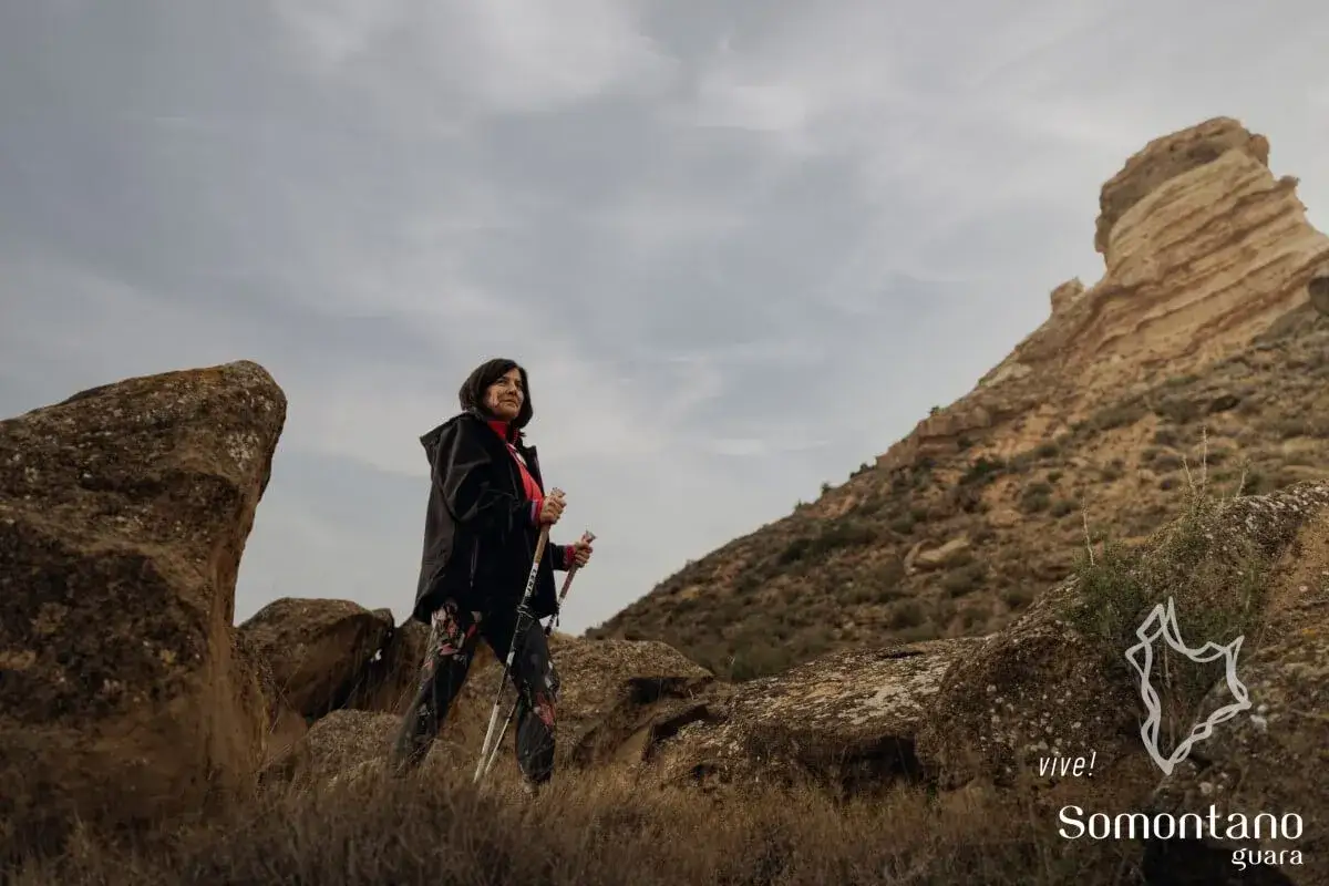 Mujer con bastones de senderismo posa en la **sierra de la Muela**, con formaciones rocosas y un cielo nublado.