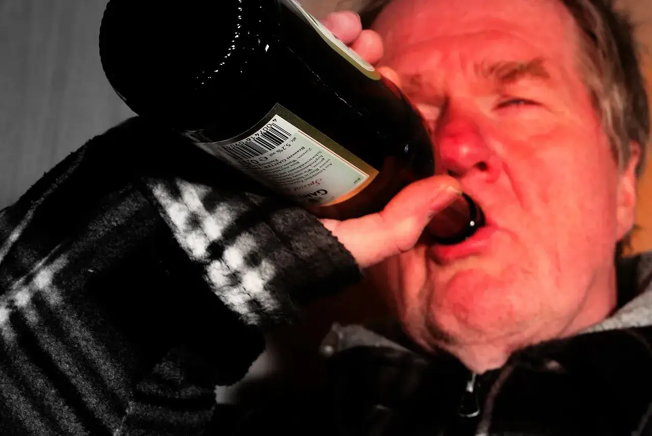 Mężczyzna z zaczerwienioną twarzą pije prosto z butelki. To obraz walki z alkoholizmem, która pochłania go coraz bardziej.