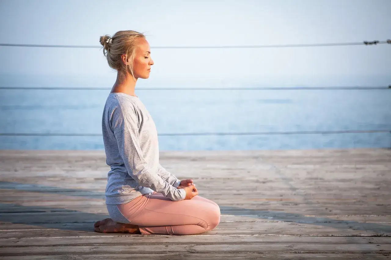Kvinna med slutna &ouml;gon sitter i meditationsst&auml;llning p&aring; en tr&auml;brygga vid havet. Hon ut&ouml;var body scanning f&ouml;r inre lugn.