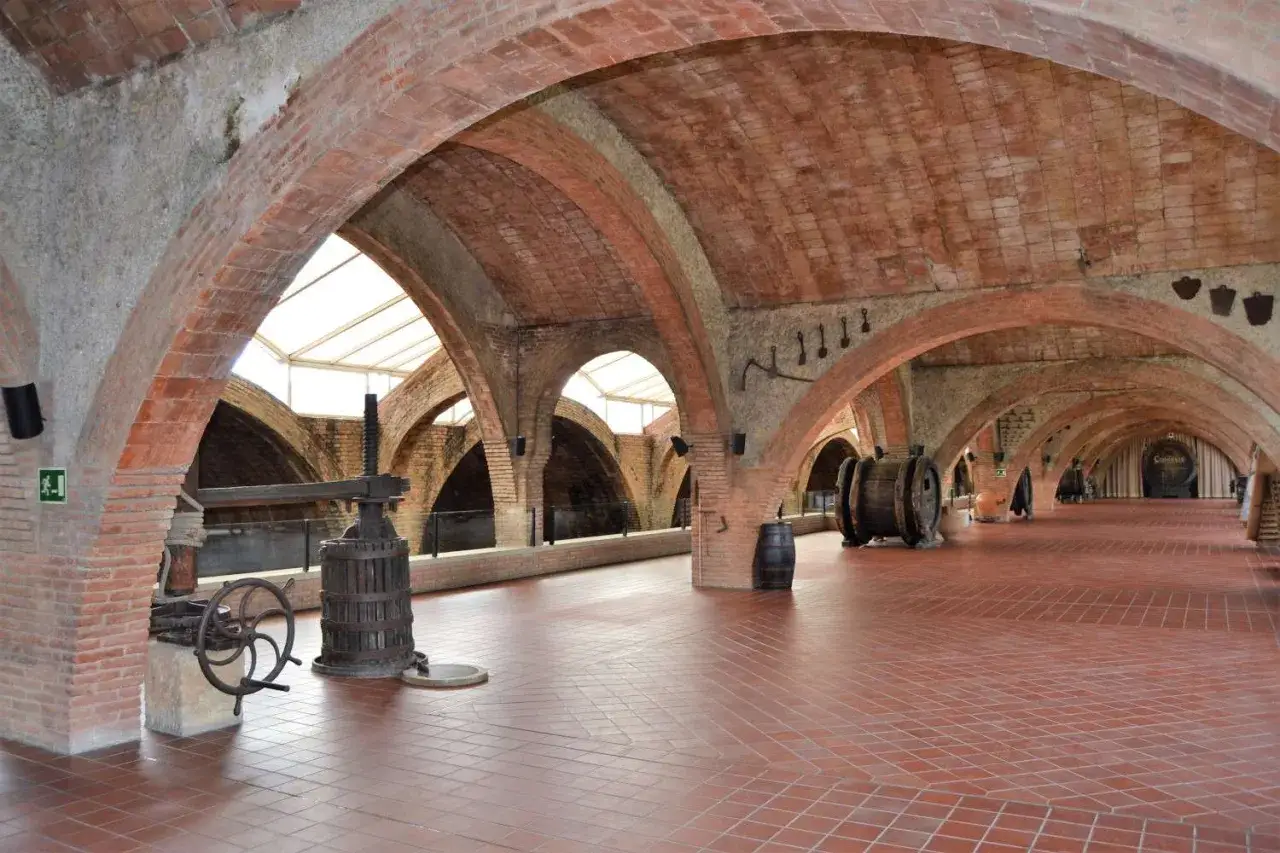 Antiguas bodegas de cava Penedès con arcos de ladrillo, prensa de vino y barricas de madera.