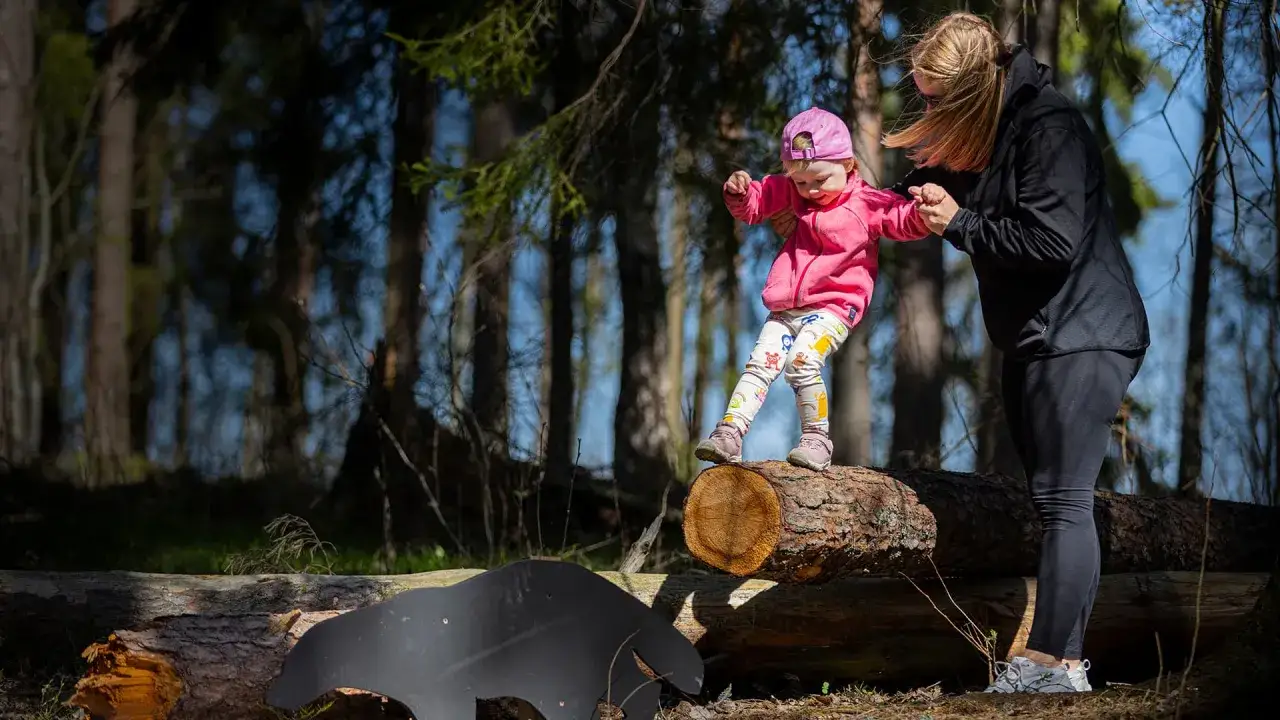 Ett litet barn i rosa jacka och m&ouml;nstrade byxor balanserar p&aring; en stock i skogen, med en vuxen som h&aring;ller i handen. &Auml;ventyrliga barn i skogen.