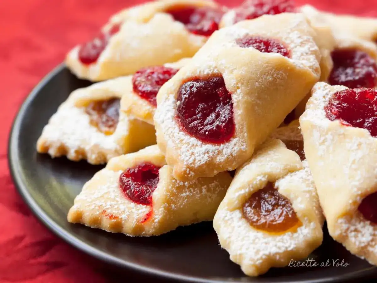 Ciastka kruche con ripieno di marmellata, spolverati di zucchero a velo, su un piatto scuro.