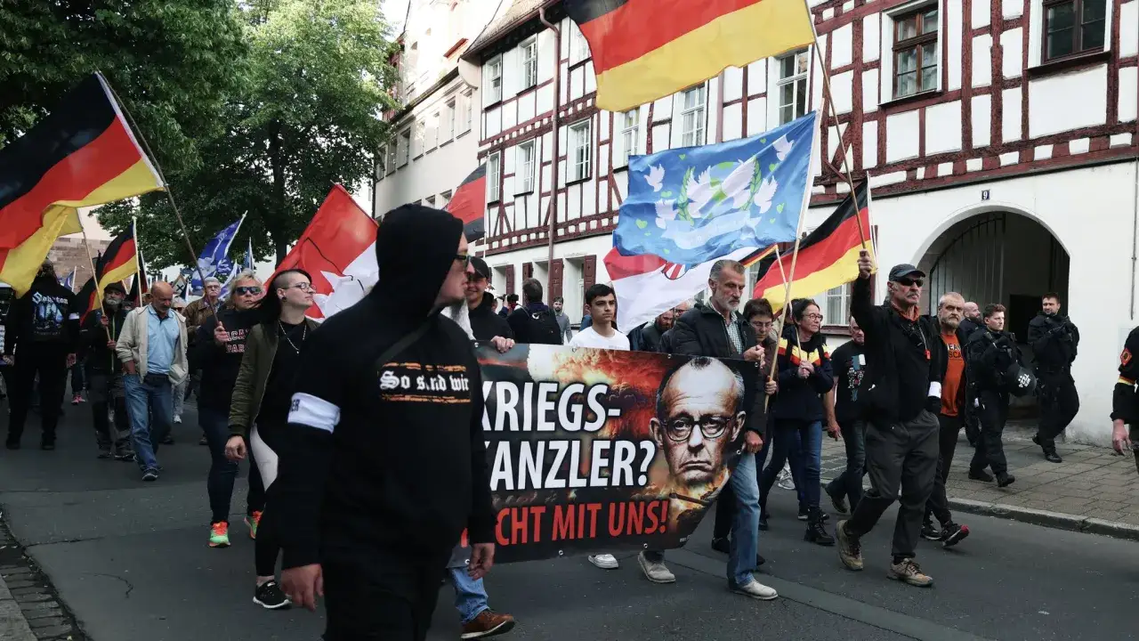 Demonstranten mit deutschen Fahnen und einem Banner, das einen Mann mit Brille zeigt. Die Botschaft auf dem Banner kritisiert einen 