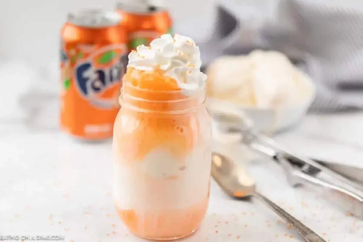Erfrischender Fanta Eis Float im Glas, garniert mit Sahne und Streuseln. Daneben stehen Fanta-Dosen und eine Schüssel mit Eis.