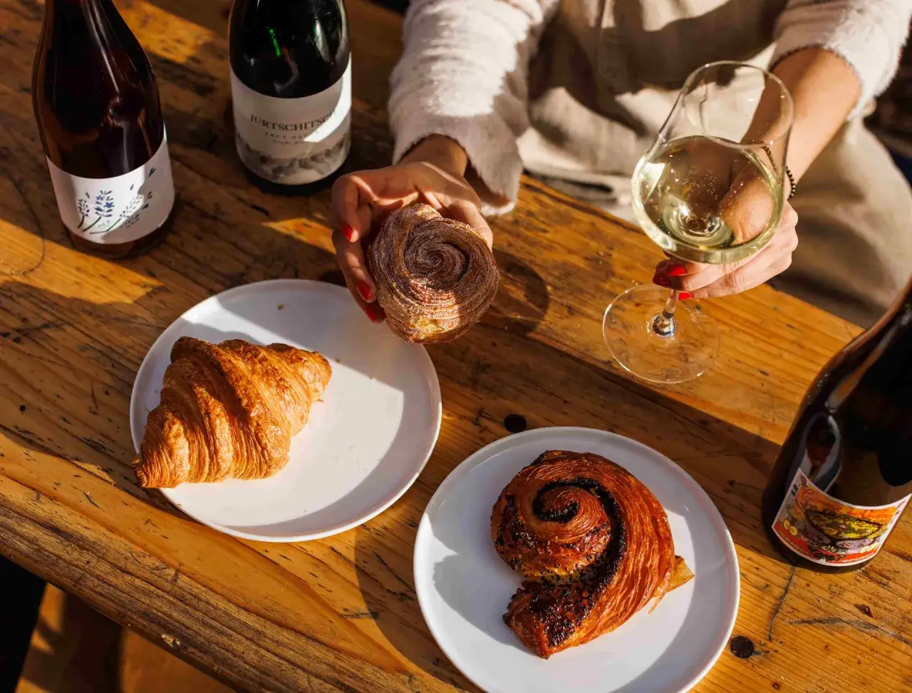 En hand h&aring;ller en sockerdr&auml;nkt bakelse bredvid en croissant och en chokladsn&auml;cka. Flera flaskor vin, varav en m&auml;rkt 