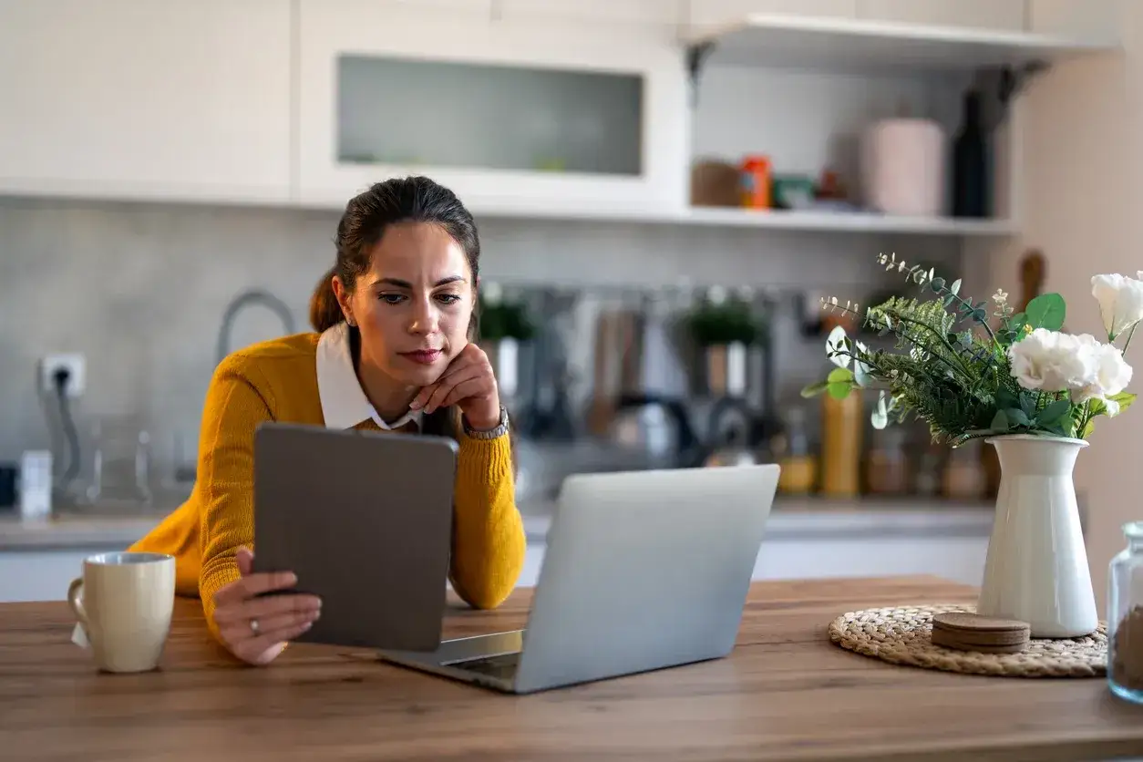 Donna studia il mutuo fino a che et&agrave;, con tablet e laptop sul tavolo della cucina.