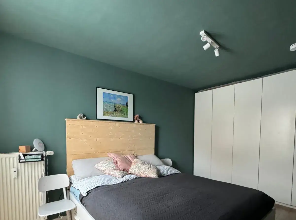 Ein Schlafzimmer mit dunklen, grünen Wänden und Decke, die in gleicher Farbe gestrichen sind. Ein Bett mit Holzkopfteil und ein weißer Schrank.