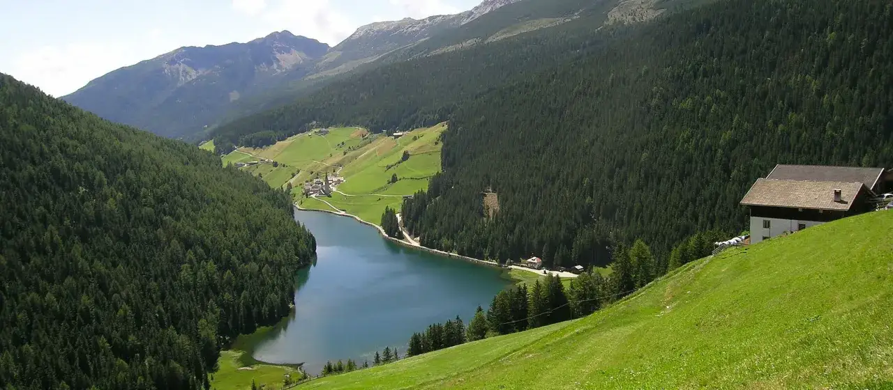 Un incantevole lago di Valdurna incastonato tra montagne boscose e verdi pascoli, con un piccolo villaggio e una casa isolata.