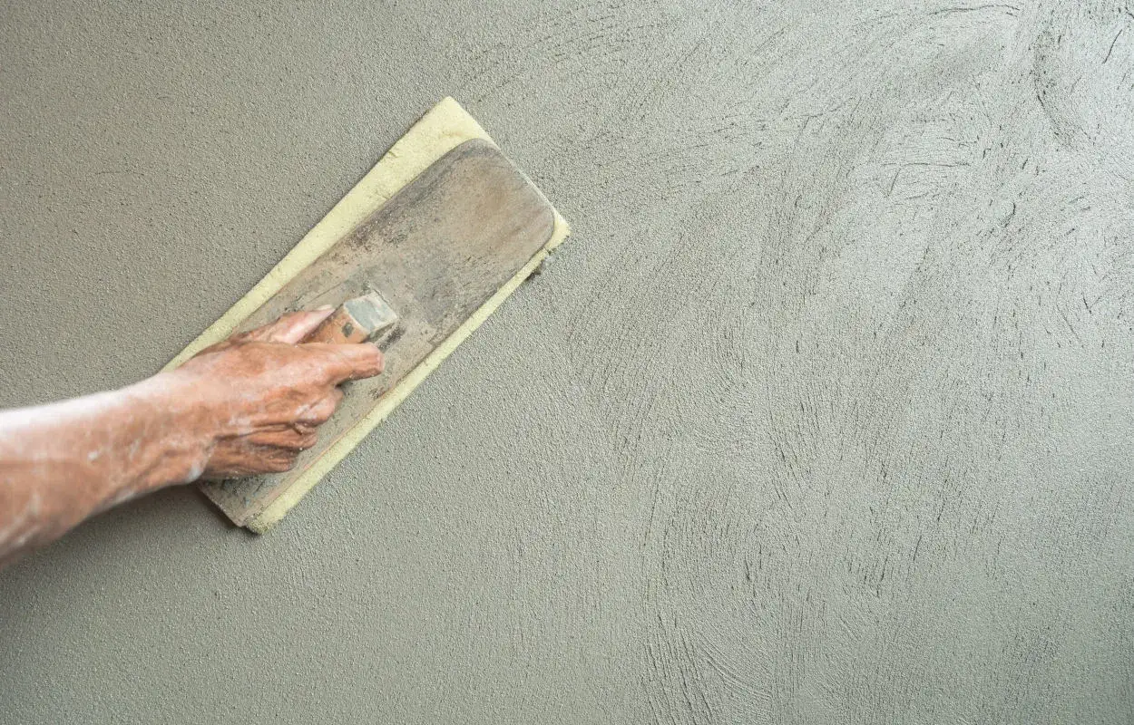 Mano che stende intonaco rustico con frattazzo, creando una texture grezza e naturale su una parete.