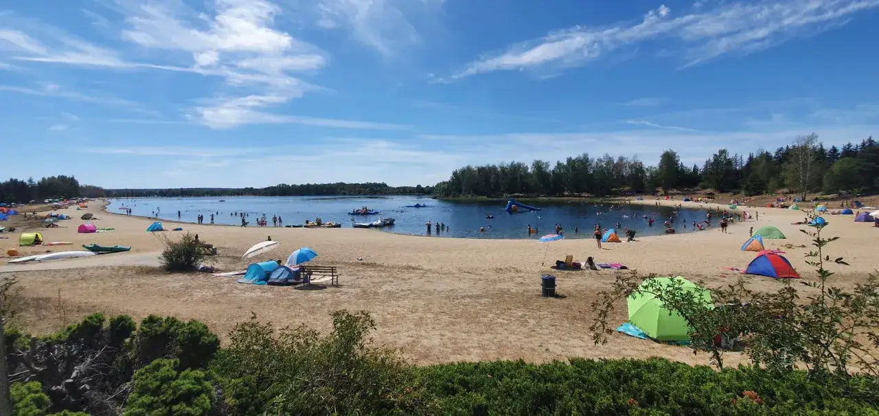 Sommerlicher Trubel am naturbad luppa: Menschen planschen im See, zelten am Sandstrand und genie&szlig;en die Sonne unter bunten Schirmen.