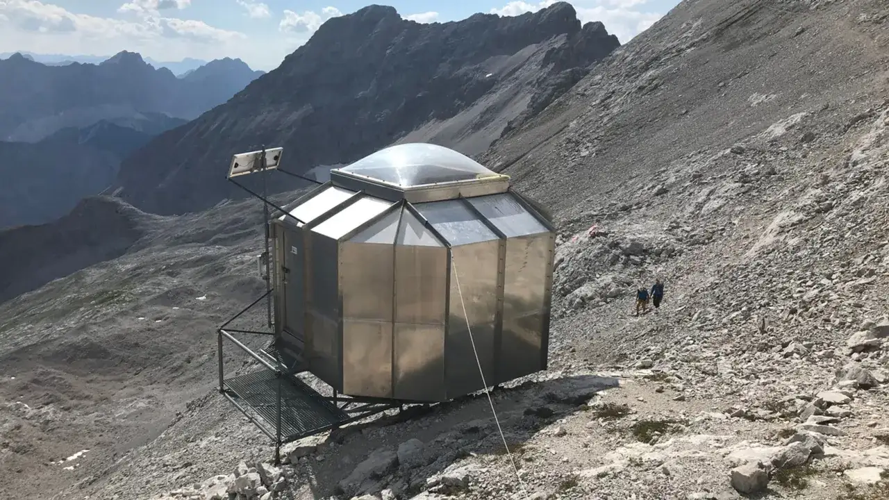 Moderne Bergh&uuml;tte mit Kuppeldach, die die Bedeutung von Biwak f&uuml;r Bergsteiger unterstreicht. Zwei Wanderer n&auml;hern sich.