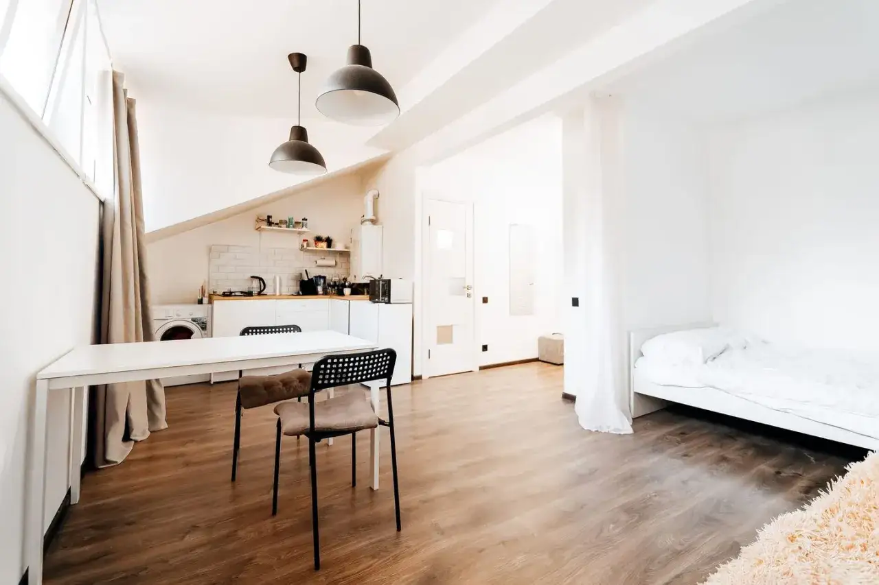 Kleiner Raum mit integriertem wohn- und schlafzimmer. Eine weiße Küchenzeile, ein Esstisch mit Stühlen und ein Bett sind sichtbar.