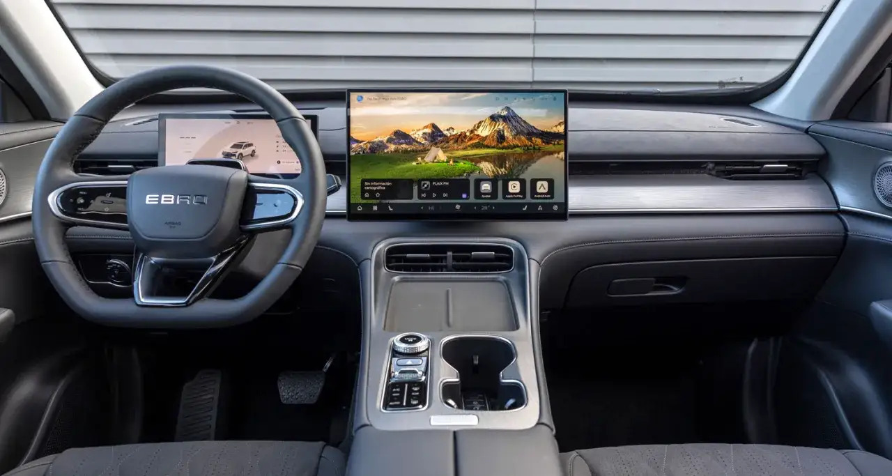 Interior del Ebro S800 PHEV con pantalla táctil central mostrando un paisaje de montaña y un volante multifunción.