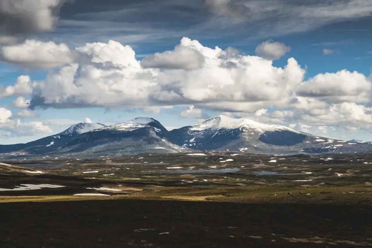 Vacker vy från en webbkamera vid Helagsfjällen. Snöklädda toppar möter en dramatisk molnhimmel över ett kuperat landskap med sjöar och grönska.