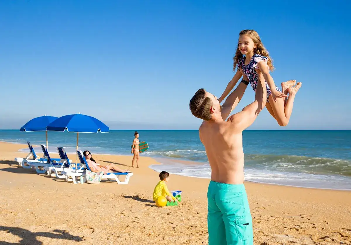 Vater hebt Tochter am Strand hoch. Familie genießt Urlaub im TUI Kids Club Barrosa Garden.