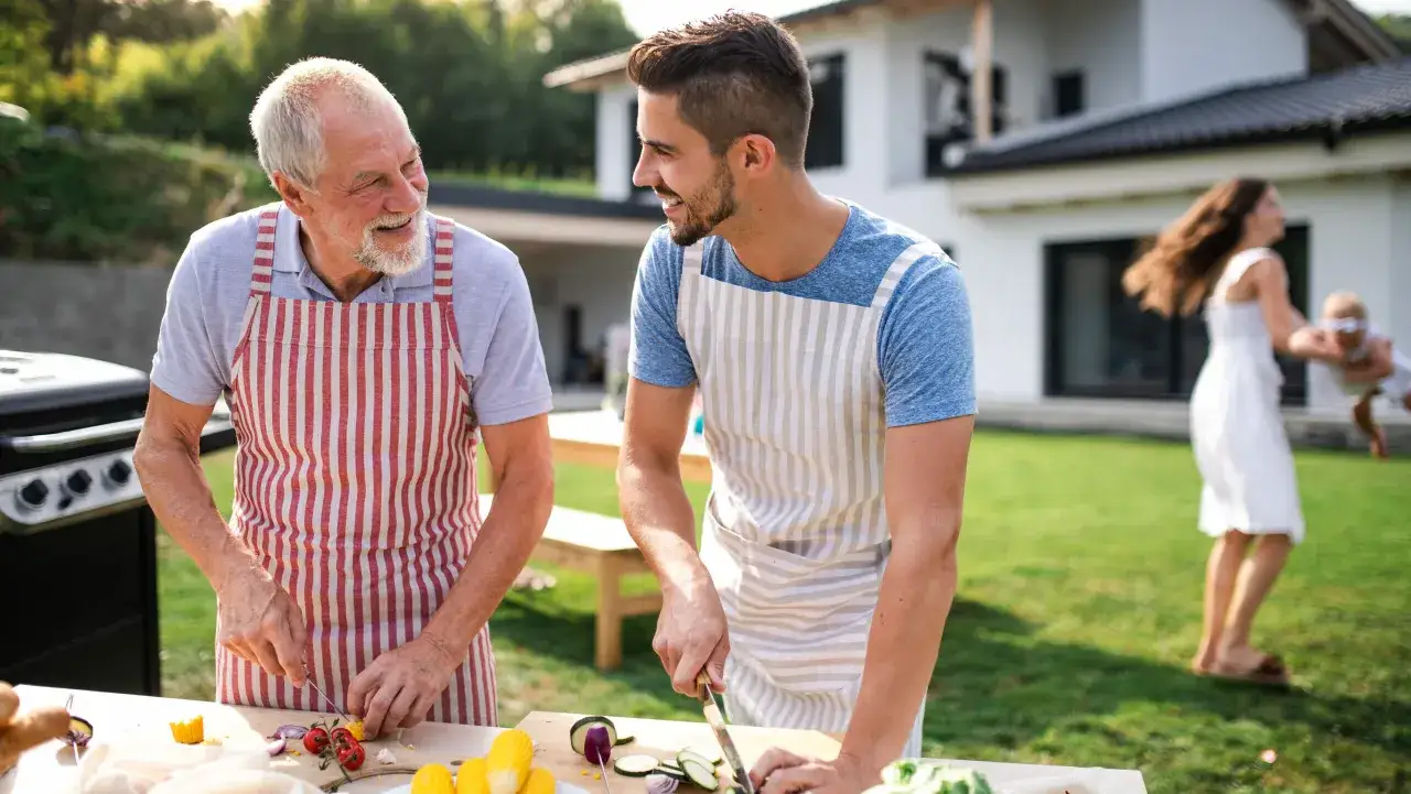 Dzień zięcia w ogrodzie. Dziadek i zięć przygotowują grill, w tle kobieta z dzieckiem.