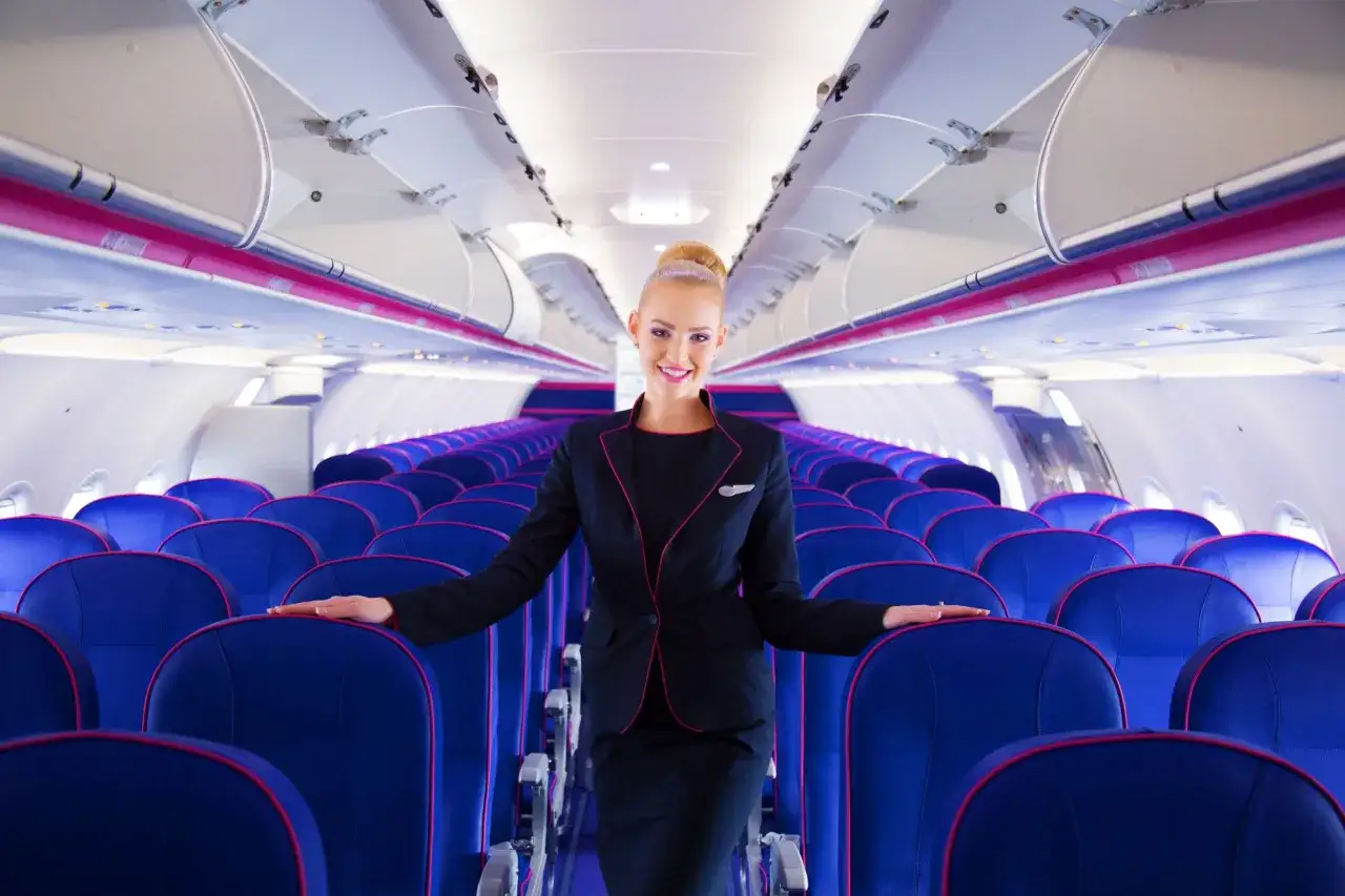 Uśmiechnięta stewardessa Wizz Air stoi między rzędami niebieskich foteli. Czy w Wizzair trzeba wybierać miejsca?