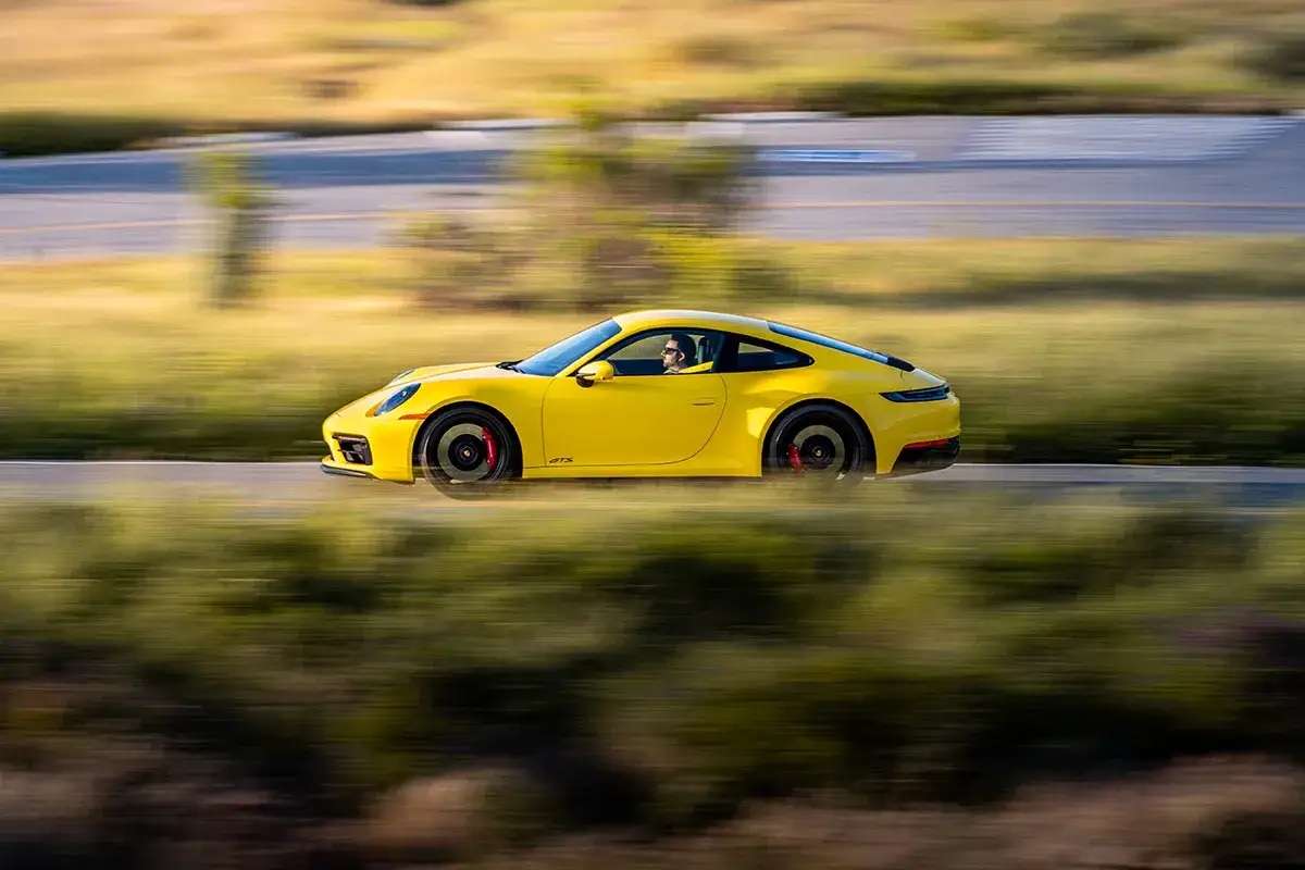 Żółty Porsche 911 Carrera GTS pędzi po krętej drodze, otoczony rozmytą zielenią i złotym światłem.