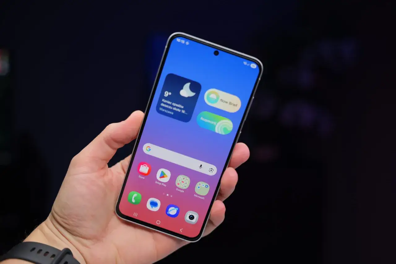 Dłoń trzyma smartfon z interfejsem One UI 8.5, wyświetlającym widżety pogody i ikony aplikacji.