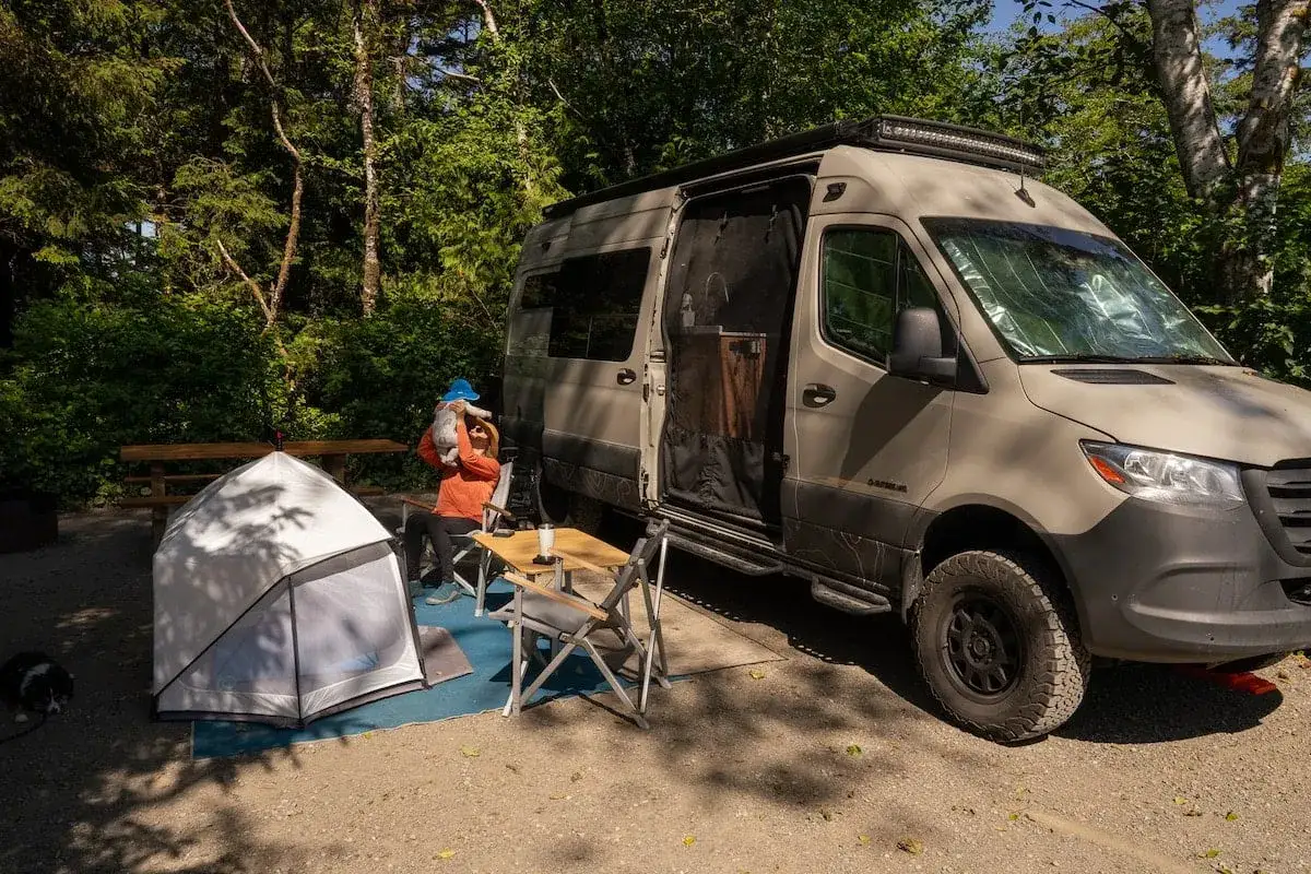 Una mujer con un gato en brazos se sienta junto a un vehículo vivienda en un campamento. Hay una tienda de campaña y una mesa plegable.