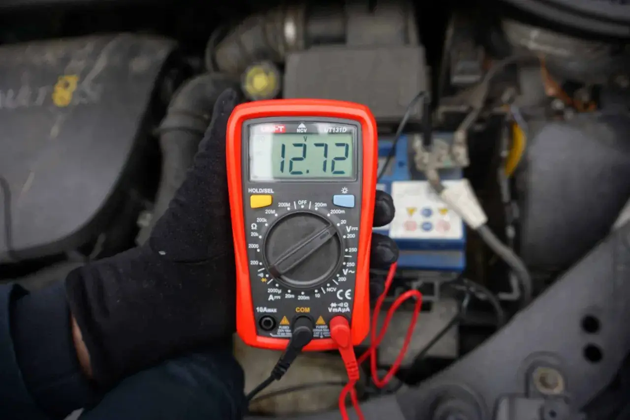 Ręka w czarnej rękawiczce trzyma multimetr, który pokazuje 12.72V. W tle widać silnik samochodu i akumulator, który może mieć migającą kontrolkę akumulatora.