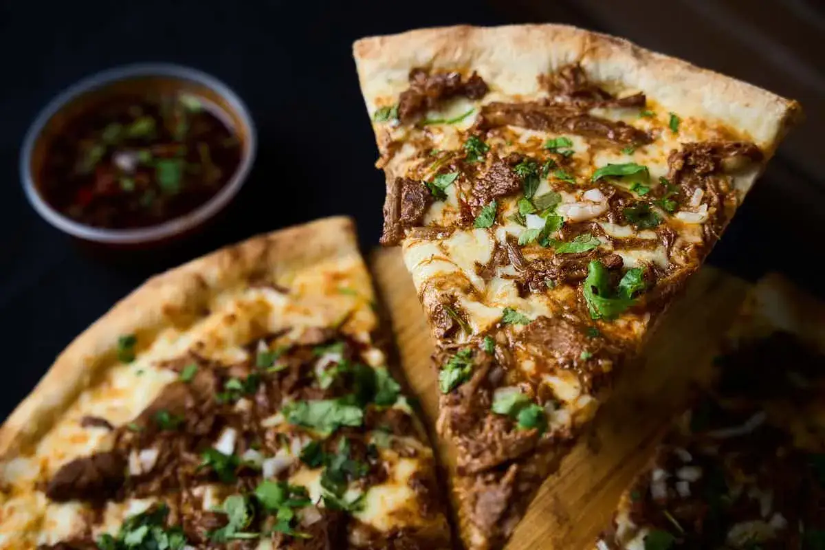 Kawałek pizzy z szarpaną wołowiną i serem, obok miseczka z sosem. Idealne dodatki do pizzy na wieczór.