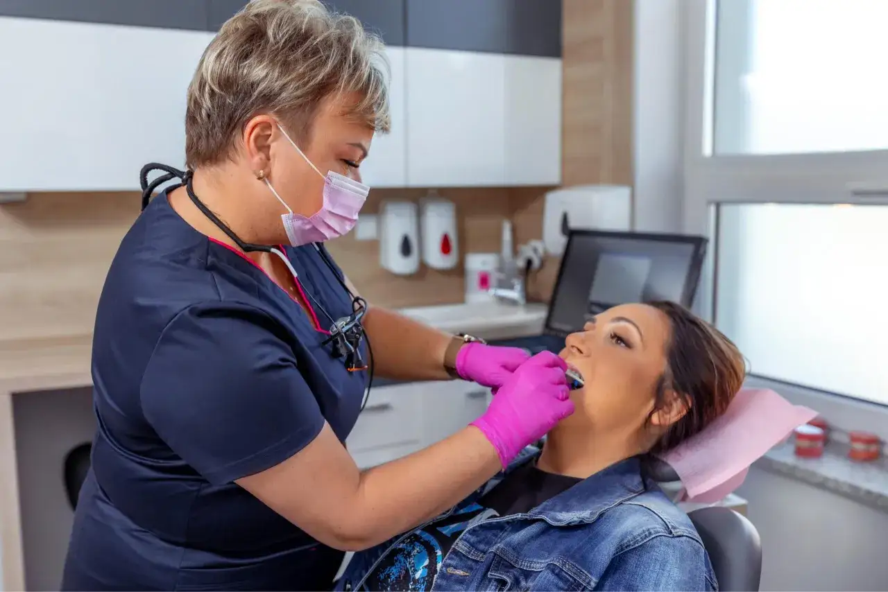 Dentystka w masce i różowych rękawiczkach bada zęby pacjentki w gabinecie.