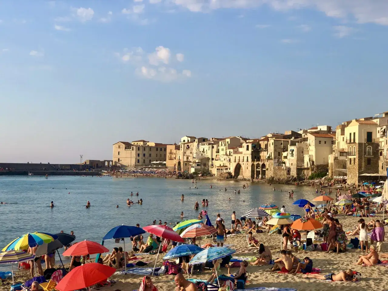 Słoneczny dzień na plaży w Cefalu, Sycylia. Tłumy ludzi odpoczywają pod parasolami, kąpią się w morzu, a w tle widać urokliwe kamienne budynki.