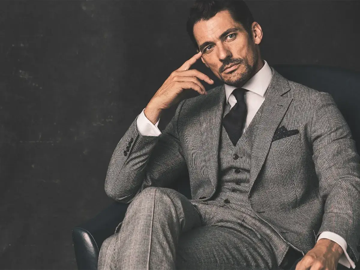 A man in a grey plaid suit, complete with a vest, white dress shirt, and dark tie, poses thoughtfully.