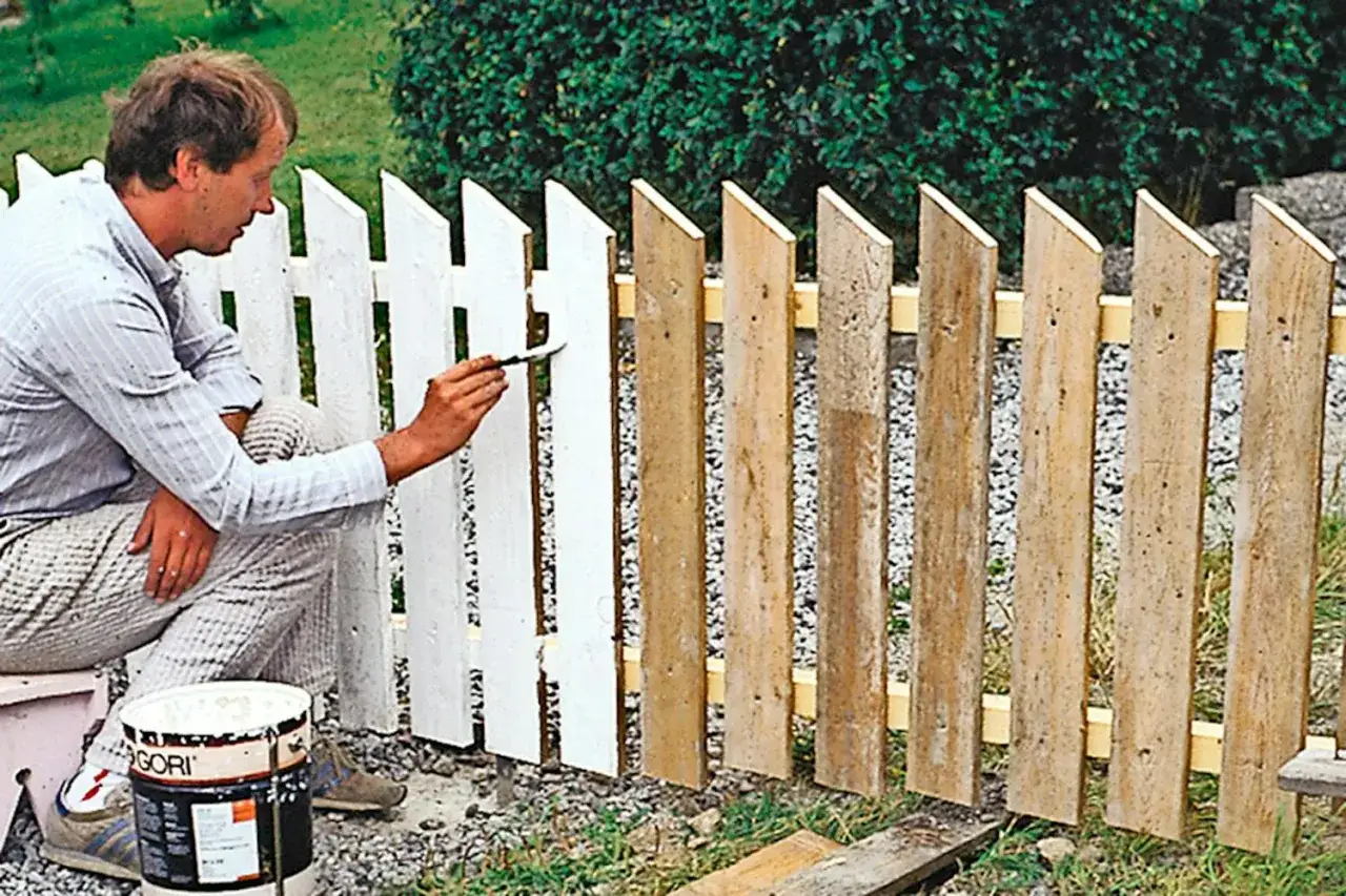 Man målar ett staket vitt. Han sitter på huk bredvid en färgburk och penslar en av träplankorna.