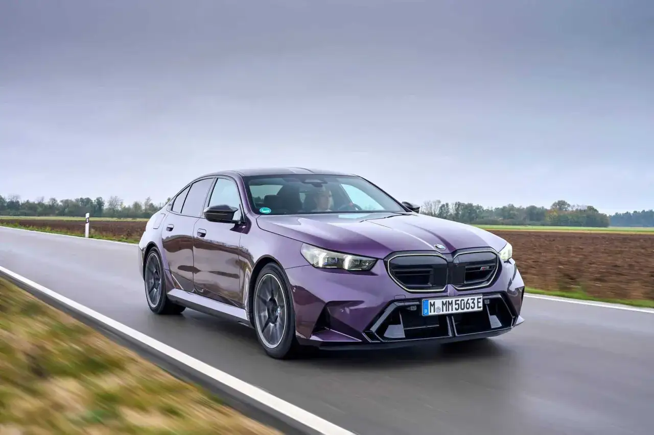 Fioletowe BMW M5 G90 pędzi po mokrej drodze, otoczone polami i drzewami.