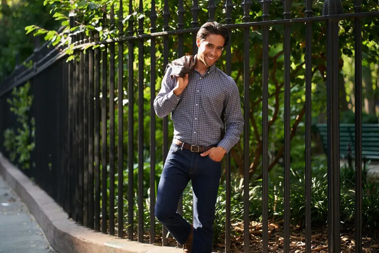 Un homme souriant porte une chemise à carreaux et une tenue jean bleu foncé, une veste en cuir sur l'épaule, appuyé contre une clôture noire.